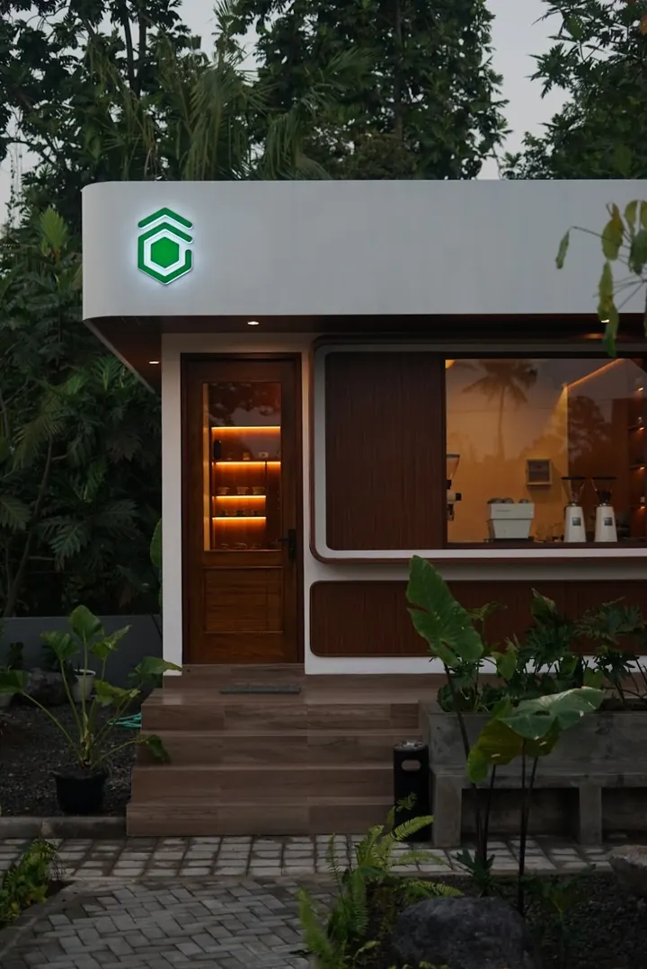 The kedai at dusk, its glowing green hexagon logo above a warm-lit timber door.