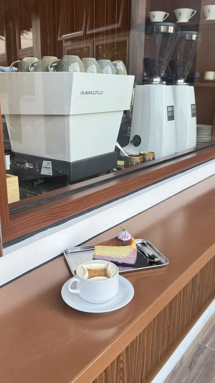 A cup of coffee and a slice of cheesecake on the timber counter by the window.
