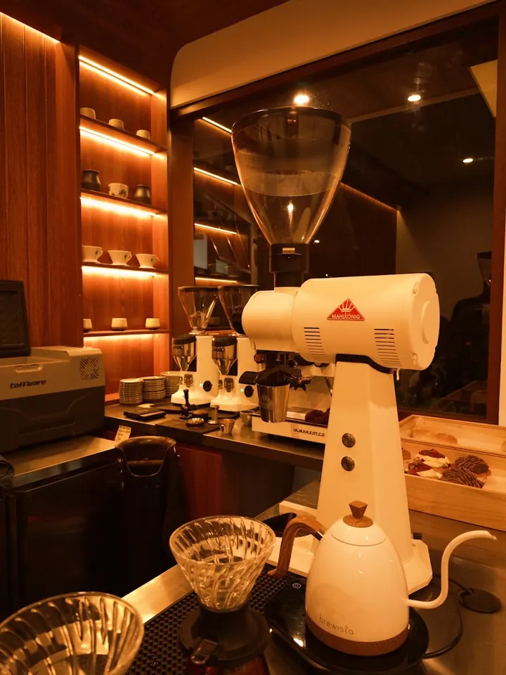Inside the bar: a white coffee grinder, gooseneck kettle and pour-over glass under warm shelf lights.