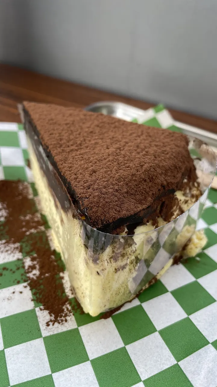 A generous slice of tiramisu dusted with cocoa on green checked paper.
